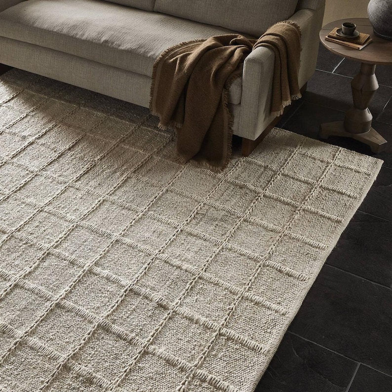 May include: A cream-colored woven rug with a grid pattern. The rug is placed under a light brown sofa with a brown throw blanket.