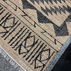 May include: A close-up of a jute rug with a black geometric pattern. The rug has a fringe border and the text "RUGS EXPORTS" is visible in the bottom right corner.