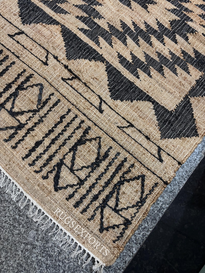 May include: A close-up of a jute rug with a black geometric pattern. The rug has a fringe border and the text "RUGS EXPORTS" is visible in the bottom right corner.