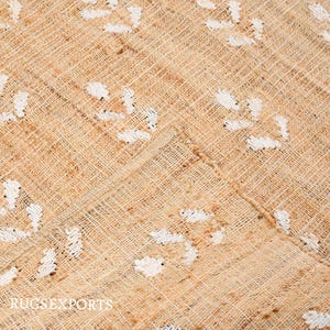May include: A close-up of a beige woven rug with a white geometric pattern. The rug has a natural, rustic look and feel.
