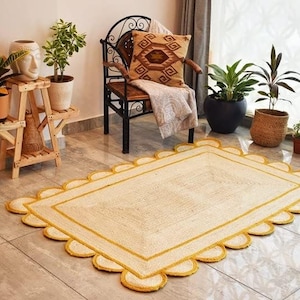 May include: A cream-colored jute rug with a yellow scalloped border. The rug is laid out on a tiled floor in a room with a chair, plants, and a wooden plant stand.