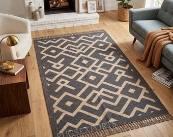 May include: A rectangular area rug featuring a dark charcoal gray background with a repeating geometric pattern in a light beige color. The rug has a flat-woven texture and is finished with short, knotted fringe along the two shorter ends.