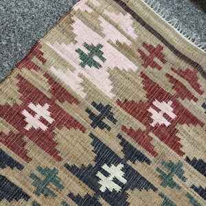 Puede incluir: Primer plano de una alfombra tejida con un patr&oacute;n geom&eacute;trico. La alfombra presenta un fondo beige con formas de diamante repetidas en rojo, rosa, verde, azul marino y blanco. El texto "RUGSEXPORTS" es visible en la esquina inferior derecha.