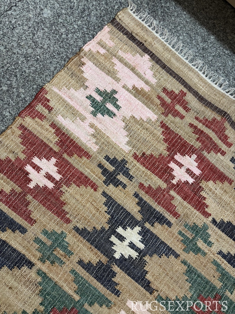 Puede incluir: Primer plano de una alfombra tejida con un patr&oacute;n geom&eacute;trico. La alfombra presenta un fondo beige con formas de diamante repetidas en rojo, rosa, verde, azul marino y blanco. El texto "RUGSEXPORTS" es visible en la esquina inferior derecha.
