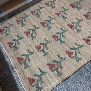 Puede incluir: Una alfombra rectangular beige con un patr&oacute;n repetido de flores rojas en forma de coraz&oacute;n y tallos verdes. La alfombra tiene un borde con flecos y el texto "RUGS EXPORTS" es visible.