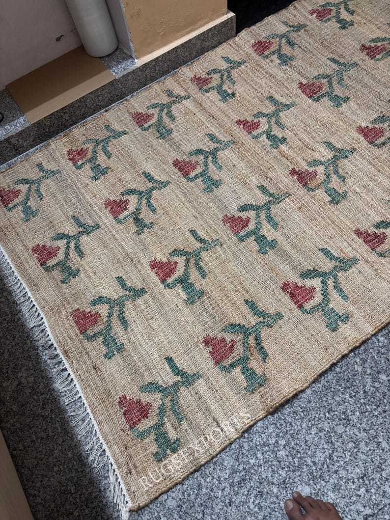 Puede incluir: Una alfombra rectangular beige con un patr&oacute;n repetido de flores rojas en forma de coraz&oacute;n y tallos verdes. La alfombra tiene un borde con flecos y el texto "RUGS EXPORTS" es visible.