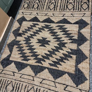 May include: Black and natural jute rug with a geometric pattern. The rug has a central diamond shape with smaller triangles and squares surrounding it. The rug has a border with a repeating pattern of lines and triangles.