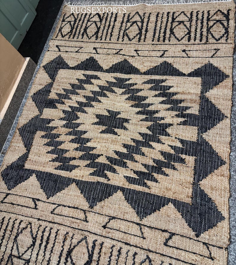 May include: Black and natural jute rug with a geometric pattern. The rug has a central diamond shape with smaller triangles and squares surrounding it. The rug has a border with a repeating pattern of lines and triangles.