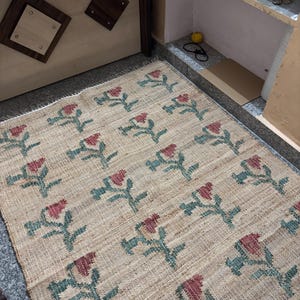 Puede incluir: Una alfombra rectangular beige con un estampado floral. Las flores son rojas con tallos y hojas verdes. La alfombra est&aacute; hecha de un material tejido y tiene un borde con flecos. El texto "RUGSEXPORTS" es visible en la parte inferior derecha.