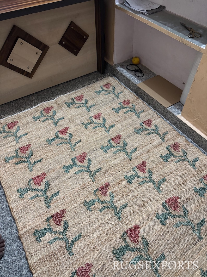 Puede incluir: Una alfombra rectangular beige con un estampado floral. Las flores son rojas con tallos y hojas verdes. La alfombra est&aacute; hecha de un material tejido y tiene un borde con flecos. El texto "RUGSEXPORTS" es visible en la parte inferior derecha.