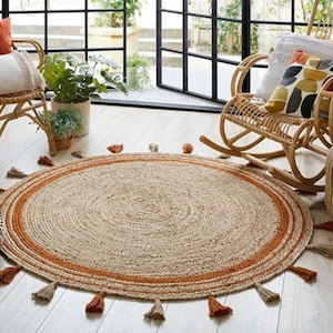 Puede incluir: Una alfombra redonda de yute de color beige natural con un borde ancho de color naranja. La alfombra tiene borlas alrededor del borde y est&aacute; colocada sobre un suelo de madera clara.