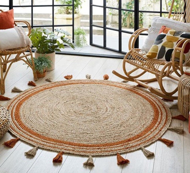 Puede incluir: Una alfombra redonda de yute de color beige natural con un borde ancho de color naranja. La alfombra tiene borlas alrededor del borde y est&aacute; colocada sobre un suelo de madera clara.