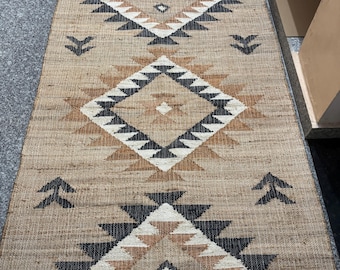 May include: A brown and black geometric patterned rug with a fringe border. The rug has a diamond shape in the center with triangles around it.