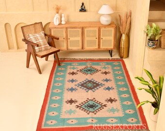 May include: A rectangular area rug featuring a beige center with repeating geometric diamond patterns in teal, brown, and black. The rug has a wide teal border with small diamond motifs and a vibrant orange outer edge with fringed ends.