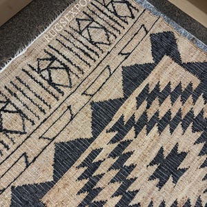 May include: A brown and black jute rug with a geometric pattern. The rug has a fringed edge and is made of natural fibers.
