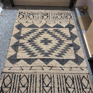 May include: Black and tan jute rug with a geometric pattern. The rug has a fringe border.
