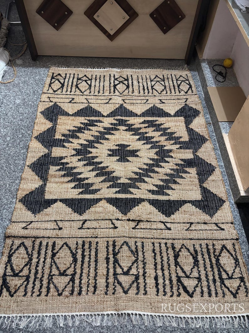 May include: Black and tan jute rug with a geometric pattern. The rug has a fringe border.