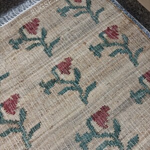 Puede incluir: Una alfombra rectangular beige con un patr&oacute;n floral repetido en rojo y verde. Las flores est&aacute;n estilizadas y dispuestas sobre la superficie de la alfombra. La alfombra tiene un borde con flecos y el texto "RUGSEXPORTS" es visible.