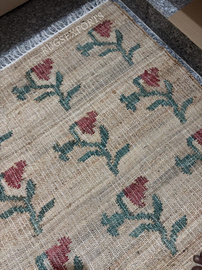 Puede incluir: Una alfombra rectangular beige con un patr&oacute;n floral repetido en rojo y verde. Las flores est&aacute;n estilizadas y dispuestas sobre la superficie de la alfombra. La alfombra tiene un borde con flecos y el texto "RUGSEXPORTS" es visible.