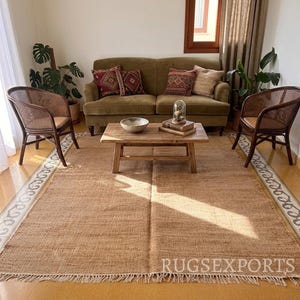 Op de afbeelding: Een groot rechthoekig geweven jute vloerkleed met een franjerand in een natuurlijke beige kleur. Het kleed ligt in een woonkamer met een houten salontafel, twee rotan stoelen en een olijfgroene bank. De tekst RUGSEXPORTS is zichtbaar.