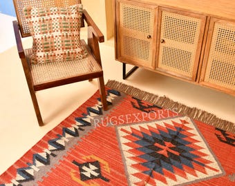 May include: A rectangular area rug featuring a red base with geometric diamond patterns in blue, white, and black, finished with fringe edges. The rug displays the text RUGSEXPORTS in white lettering across the center of the woven surface.