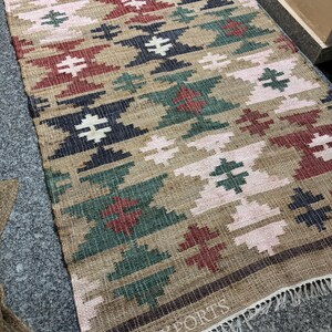 Puede incluir: Una alfombra rectangular tejida con un patr&oacute;n geom&eacute;trico en tonos beige, rojo, verde, azul marino y rosa. La alfombra tiene flecos en el borde inferior y el texto "RUGSEXPORTS" es visible.