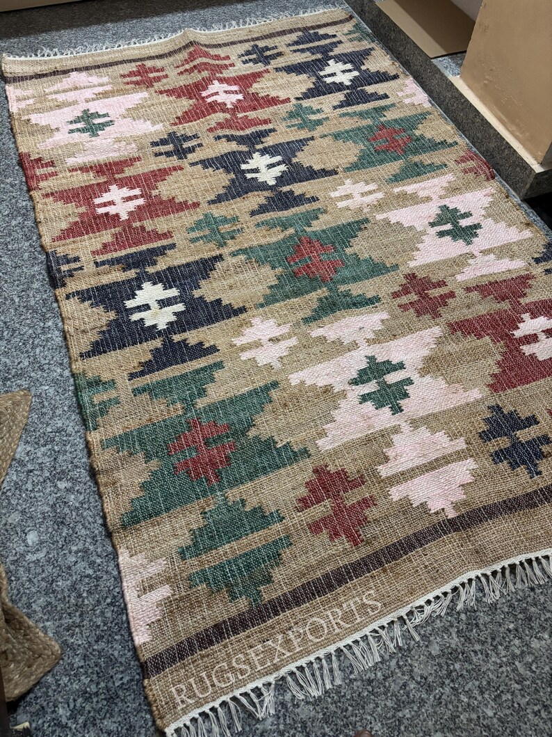 Puede incluir: Una alfombra rectangular tejida con un patr&oacute;n geom&eacute;trico en tonos beige, rojo, verde, azul marino y rosa. La alfombra tiene flecos en el borde inferior y el texto "RUGSEXPORTS" es visible.