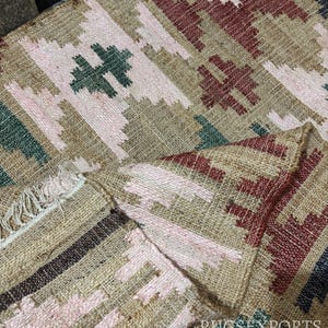 Puede incluir: Alfombra tejida con un patr&oacute;n geom&eacute;trico en tonos beige, rosa, rojo, verde y negro. La alfombra presenta un dise&ntilde;o repetitivo de formas de diamante y motivos en forma de cruz, con un borde con flecos.