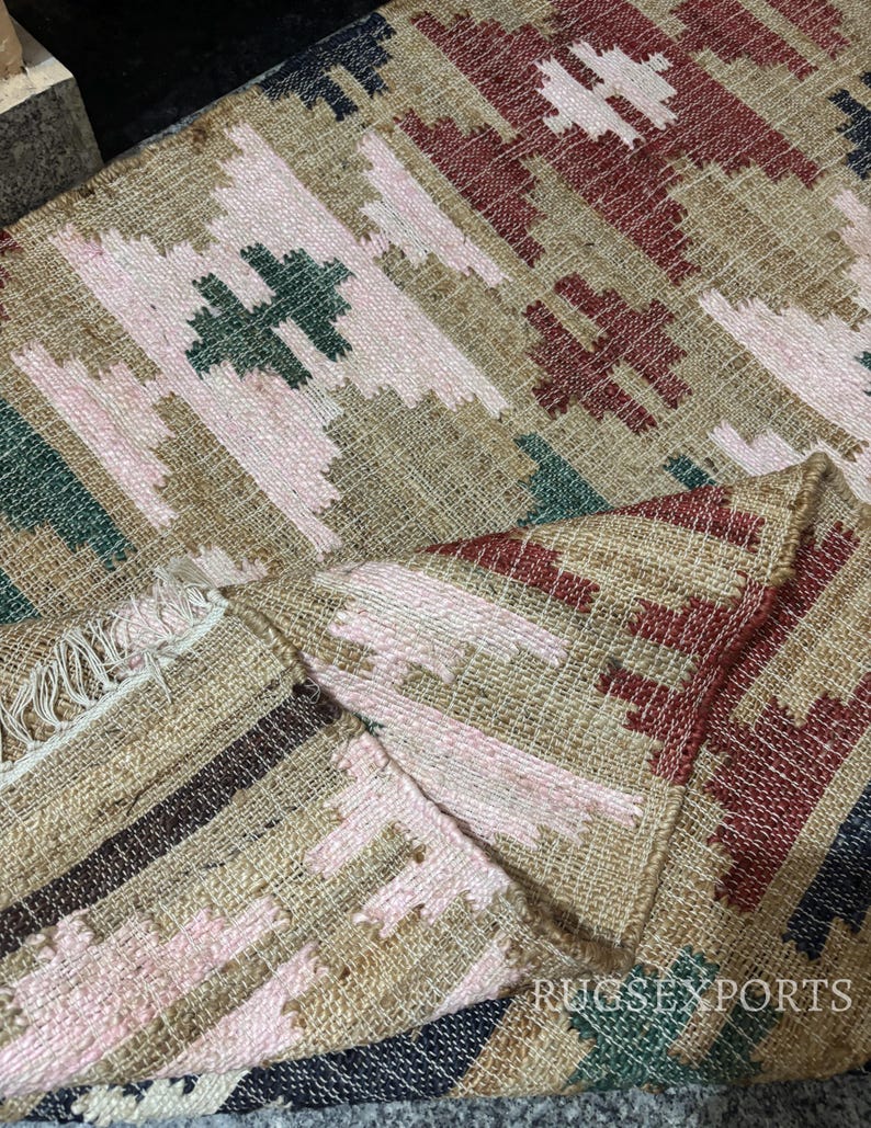 Puede incluir: Alfombra tejida con un patr&oacute;n geom&eacute;trico en tonos beige, rosa, rojo, verde y negro. La alfombra presenta un dise&ntilde;o repetitivo de formas de diamante y motivos en forma de cruz, con un borde con flecos.