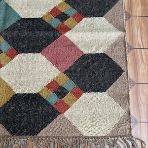 Pu&ograve; includere: Un tappeto geometrico con un motivo a rombi in vari colori. Il tappeto presenta una combinazione di nero, bianco, rosa, verde acqua, giallo senape e marrone. Il tappeto ha una frangia lungo il bordo inferiore.