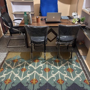 May include: A home office with a wooden desk, two chairs, a laptop computer, and a patterned rug. The desk has two water bottles, a pen holder, and a laptop computer on it. The rug is blue, green, and yellow with a geometric pattern.