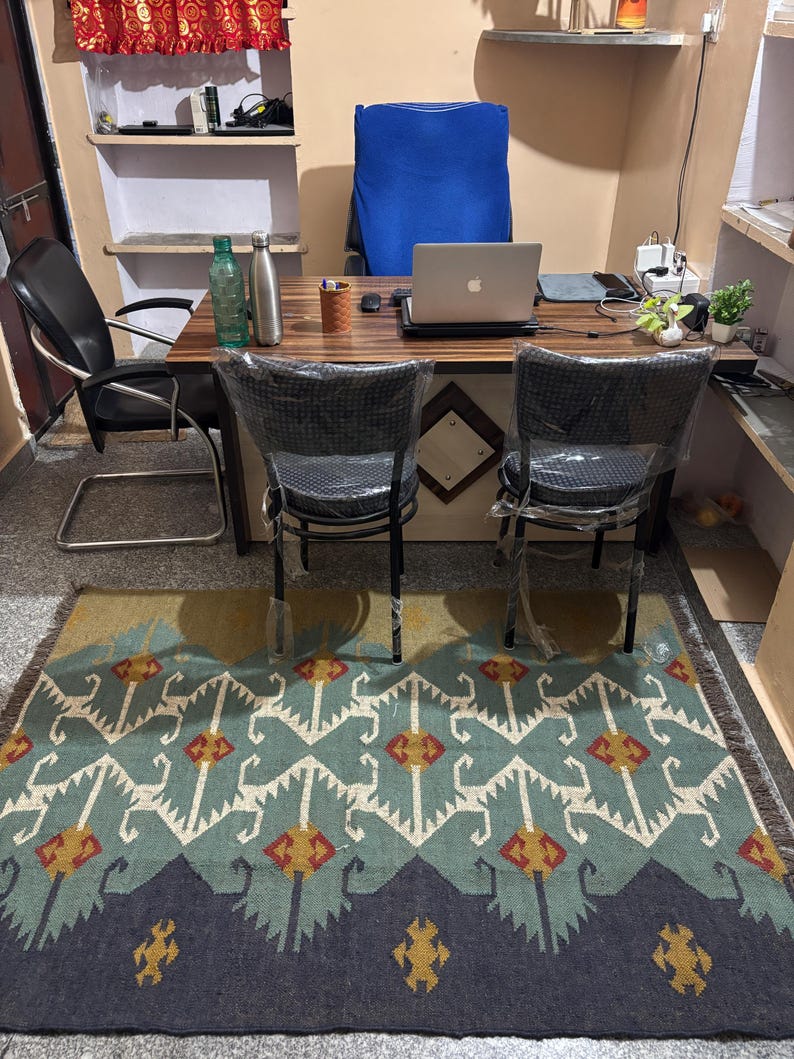 May include: A home office with a wooden desk, two chairs, a laptop computer, and a patterned rug. The desk has two water bottles, a pen holder, and a laptop computer on it. The rug is blue, green, and yellow with a geometric pattern.