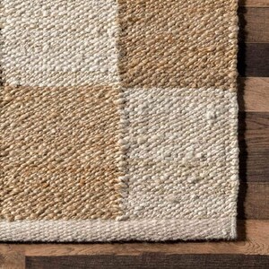 May include: Close-up of a woven rug with a checkered pattern in beige and white. The rug has a textured surface and a fringe border.