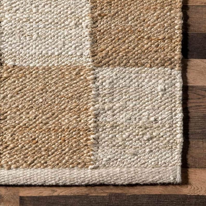 May include: Close-up of a woven rug with a checkered pattern in beige and white. The rug has a textured surface and a fringe border.