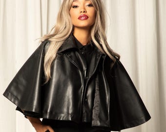 May include: A black leather cape jacket with a collared neckline and button closure. The model is wearing black pants and black combat boots. The background is a white curtain.