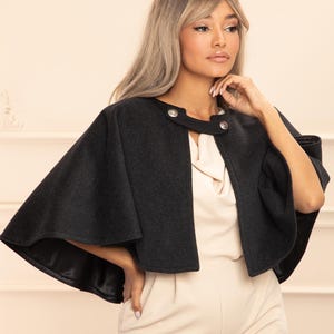 Può includere: Un mantello grigio scuro con scollatura rotonda e due bottoni argentati. Il mantello ha un design svasato ed è indossato sopra un top e pantaloni color crema. La fodera del mantello è nera. L'outfit è un insieme elegante.