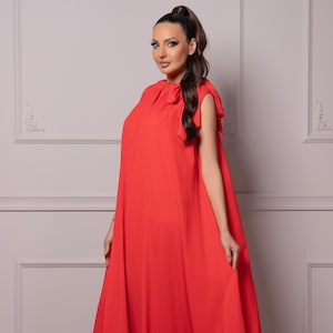 Könnte beinhalten: Ein leuchtend rotes, bodenlanges Kleid mit fließender Silhouette. Das Kleid hat einen hohen Ausschnitt mit Schleifen an den Schultern. Das Model trägt schwarze Absätze.