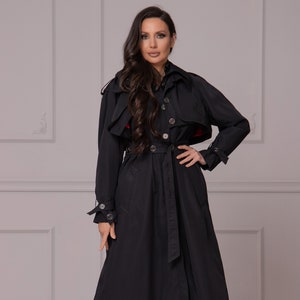 Op de afbeelding: Een zwarte trenchcoat met een riem en een capuchon. De jas heeft een dubbele rij knopen en zilveren knopen.