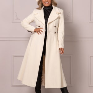 May include: A long white coat with a double-breasted closure and a flared skirt. The coat has brown buttons and is worn over a black turtleneck top and black trousers. The person is wearing a white hat with a brown button.