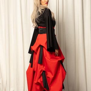 May include: A woman wearing a black top with long sleeves and a red skirt with a ruffled design. The skirt has black ties at the waist and on the ruffles. She is wearing black heels.