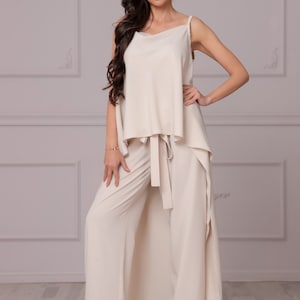 May include: A cream-colored two-piece outfit with a wide-leg pant and a loose-fitting top. The top has a draped front and a tie at the waist. The model is wearing red heels.