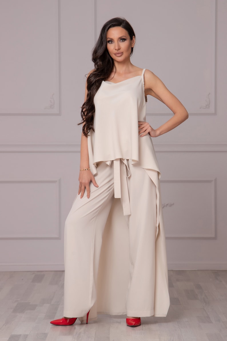 May include: A cream-colored two-piece outfit with a wide-leg pant and a loose-fitting top. The top has a draped front and a tie at the waist. The model is wearing red heels.