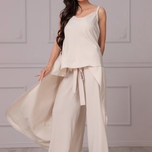 May include: A woman wearing a cream-colored pantsuit with a flowing top and wide-leg pants. The pants have a sash tie at the waist. She is wearing red high heels.