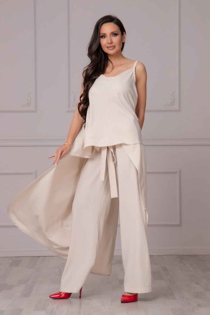May include: A woman wearing a cream-colored pantsuit with a flowing top and wide-leg pants. The pants have a sash tie at the waist. She is wearing red high heels.