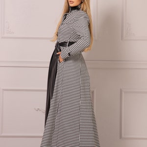 May include: A long black and white houndstooth coat with a black leather belt. The coat has a high collar and long sleeves.