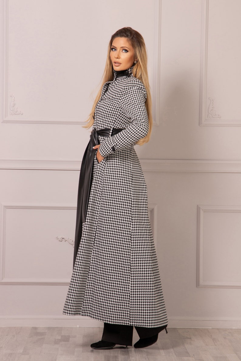 May include: A long black and white houndstooth coat with a black leather belt. The coat has a high collar and long sleeves.