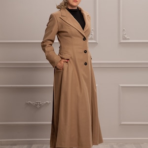 K&ouml;nnte beinhalten: Ein langer, camel-farbener Mantel mit klassischem Kragen und Knopfleiste. Der Mantel hat einen taillierten Oberk&ouml;rper und einen flie&szlig;enden Rock. Das Model tr&auml;gt rote High Heels.