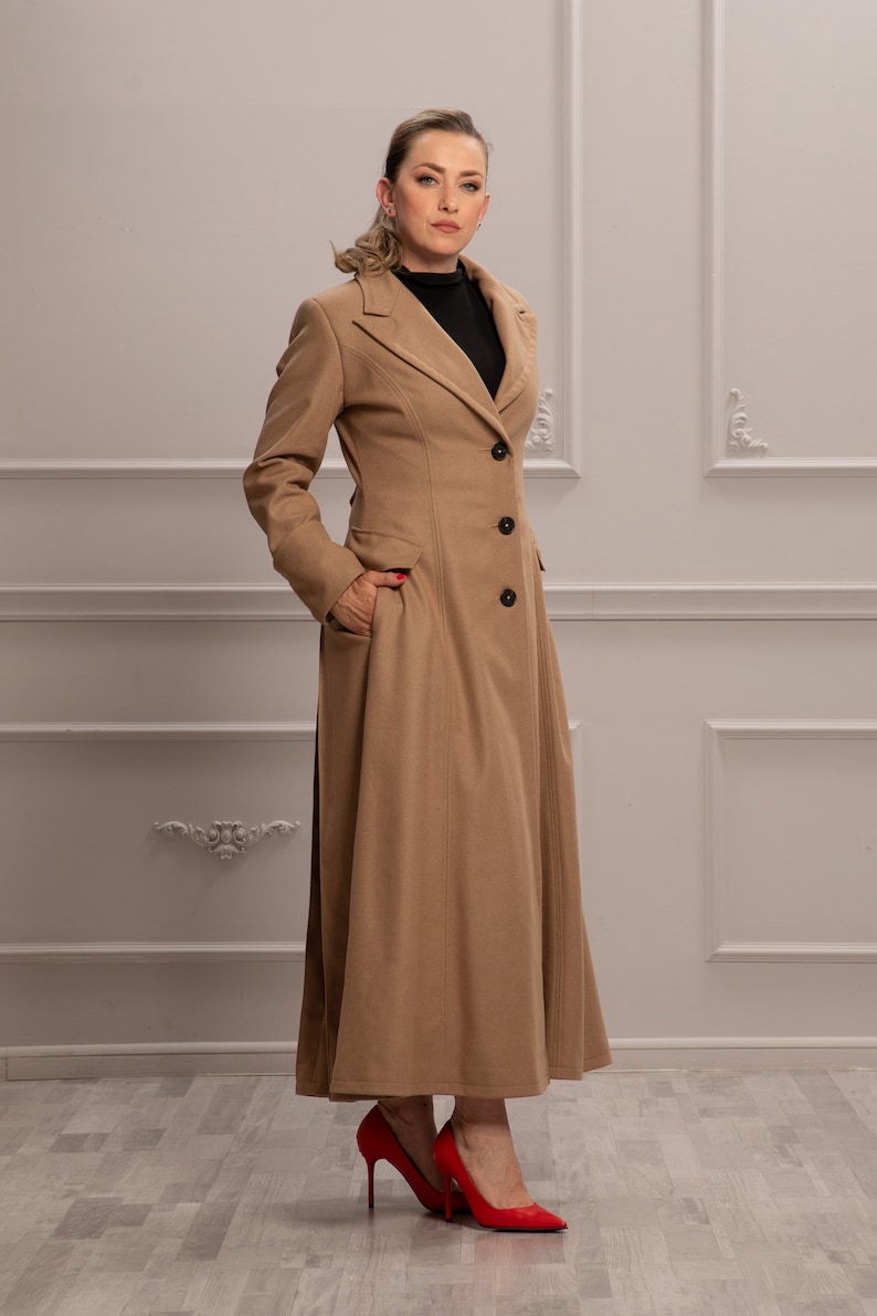 K&ouml;nnte beinhalten: Ein langer, camel-farbener Mantel mit klassischem Kragen und Knopfleiste. Der Mantel hat einen taillierten Oberk&ouml;rper und einen flie&szlig;enden Rock. Das Model tr&auml;gt rote High Heels.