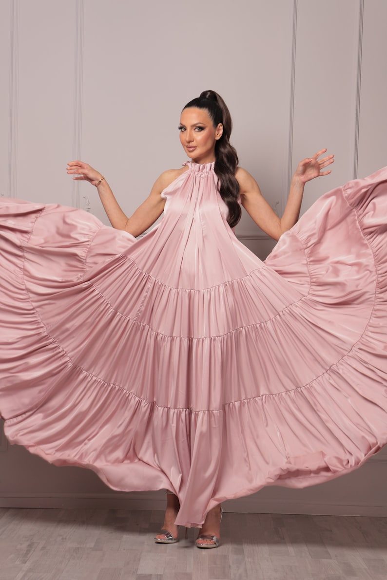 K&ouml;nnte beinhalten: Eine Frau tr&auml;gt ein langes, flie&szlig;endes rosa Kleid mit mehreren Lagen. Das Kleid ist aus seidenartigem Material und hat einen Halterneck. Die Frau steht auf einem hellen Holzboden mit einer wei&szlig;en Wand im Hintergrund.