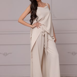 May include: A woman wears a beige pantsuit with a wide-leg, flowing pant style. The top is a sleeveless, draped design with a cowl neck. She is wearing red heels.